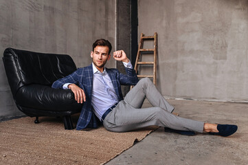 A man in a blue blazer relaxes on a chair, legs stretched out, posing in a modern interior with soft diffused lighting.