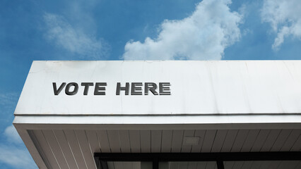 "Vote Here" word signage on a public building under a blue sky, symbolizing democracy, elections, civic duty, political participation, and the electoral process