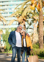 Vertical. Smiling senior couple walking arm in arm through a sunny park, sharing laughter and affection. Casual denim style, palm trees in the background, relaxed stroll on a bright day.