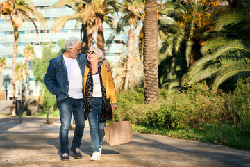 Smiling senior couple walking arm in arm through a sunny park, sharing laughter and affection. Casual denim style, palm trees in the background, relaxed stroll on a bright day.