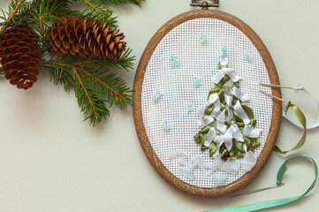 Making handmade Christmas gifts. An embroidered Christmas tree with satin and nylon ribbons on white canvas in hoop and craft accessories on table with fir branches and cones. Flat lay, copy space