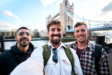 Three cheerful friends take a selfie by Tower Bridge in London, backpacks on and city views behind them. A candid travel moment with bright smiles beside the River Thames.