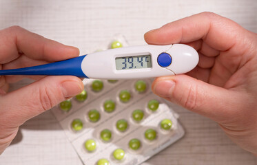 Holding a digital thermometer showing a high body temperature of 39.4 degrees Celsius next to blisters of green pills.