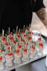 A catered silver tray filled with small glasses of shrimp cocktail topped with strawberry halves and rosemary sprigs, held by a person in black