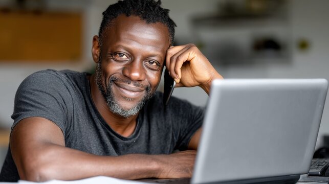 A smiling African American man sits at a table with a laptop. He is talking on his cell phone and looking at the camera - Powered by Adobe