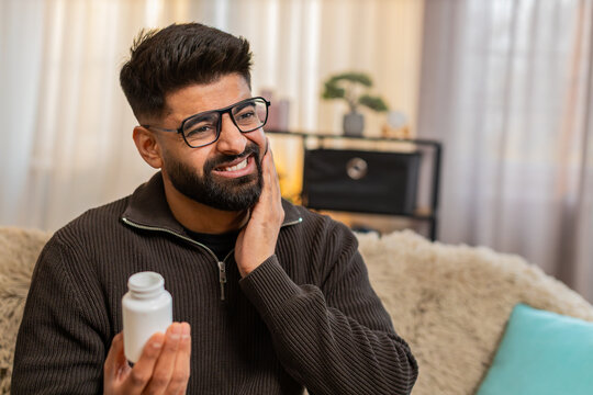 Indian man at home holds cheek with painful face looking for painkillers due to strong dental discomfort. Arabian guy on sofa reacts to toothache feeling unwell and stressed by aching health condition