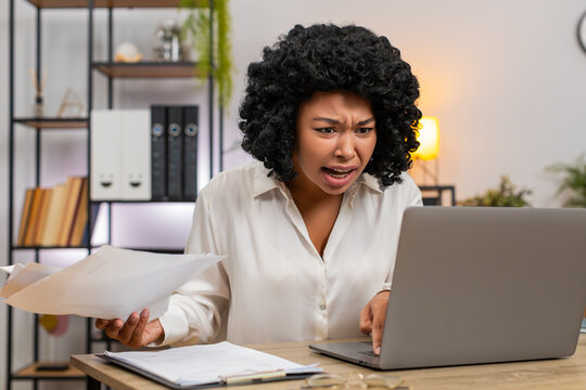 African American businesswoman at home office sees laptop crash glitch panics tossing papers urgently. Freelancer girl at table hits power button reboot hoping data safe returns and virus cleared soon