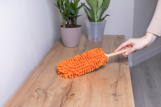 Woman removing dust from the wooden surface with an orange microfiber duster. Concept of dust removal, home cleaning, hygiene, surface care. - Powered by Adobe