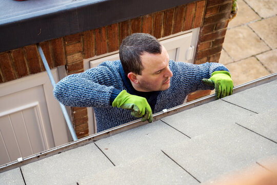 Man on a ladder cleaning fallen autumn leaves and moss from house roof gutter. Home maintenance, gutter cleaning