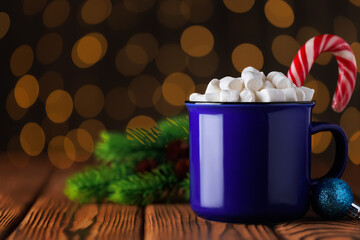 Blue cup of hot cocoa with marshmallows and candy cane on wooden table against background of bokeh glowing garland lights