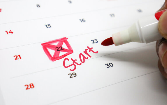 A hand uses a red marker to write Start and highlight a specific date on a calendar, symbolizing a new project, plan, or event