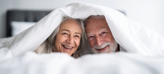 The senior couple snuggled under a white duvet smiling warmly in a bright bedroom morning
