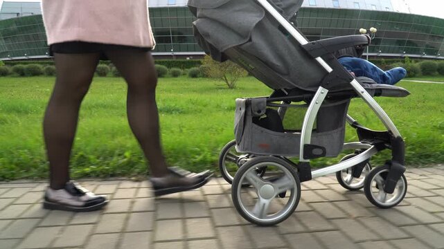 mothers legs in black tights and shoes walking beside a modern grey stroller on a paved path, baby visible inside, green park and glass building background