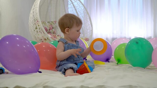 adorable toddler in denim overalls playing with colorful stacking toys on a white bed surrounded by festive balloons, happy child enjoying indoor playtime or birthday celebration