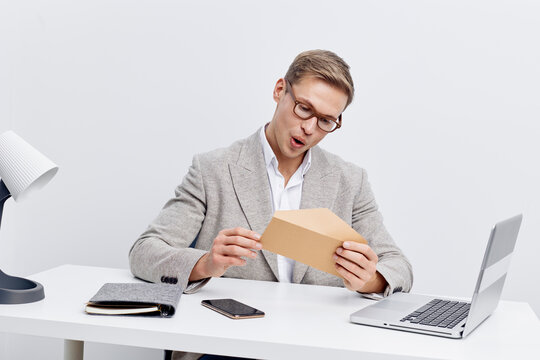 Businessman opening envelope at workspace with laptop and smartphone, professional attire, surprised expression, modern office environment, neutral background, concept: business communication and