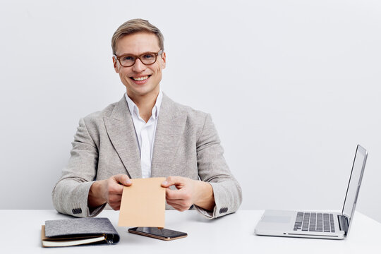 Smiling mature man in glasses and light grey blazer sitting at desk with electronic devices and notepad, holding a piece of paper, studio shot on plain background. Professional, business, office