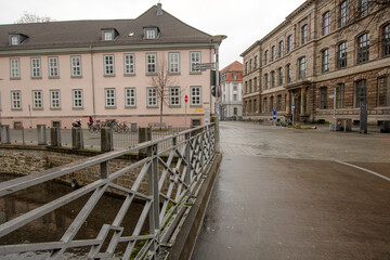 Obraz premium A simple bridge crosses a calm city canal, framed by historic façades and an open square, captured in clear urban space under soft grey daylight.
