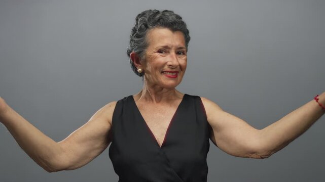 Senior woman with grey hair smiling and gesturing expressively in a sleeveless black dress against a plain grey background.