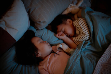 Asian girl and boy sleeping side by side under blanket in bed, both resting with stuffed animals, peaceful expressions, evening home setting, siblings sharing bedtime