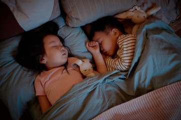Asian girl child and Asian boy child sleeping side by side in bed under blanket, both resting with stuffed animals, peaceful expressions, spending evening at home together