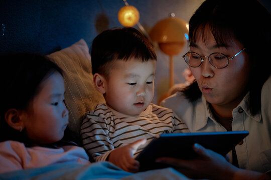 Asian woman sitting with two children in bed, holding digital tablet, children watching screen closely, woman engaging with kids during evening family time