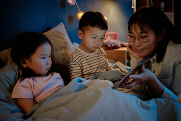 Asian woman sitting on bed showing digital tablet to girl child and boy child, children watching screen attentively during evening family time at home