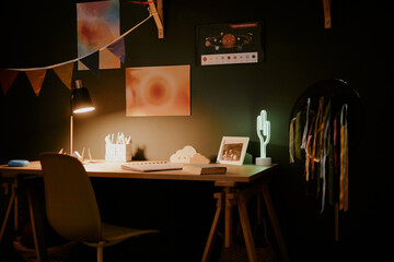 Modern home workspace featuring wooden desk with illuminated table lamp, cactus shaped neon light, framed photo, art supplies, wall art and colorful hanging ribbons visible in background