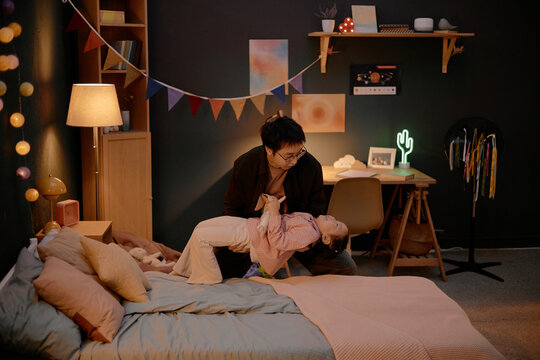 Asian middle aged man playing with Asian child in bedroom, father lifting daughter and smiling, spending evening together at home, cozy family interaction, warm indoor lighting