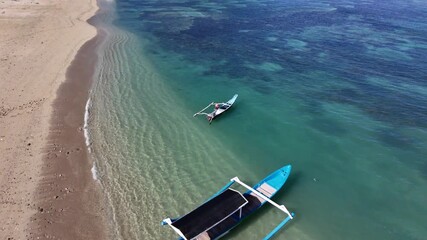 Aerial top down view of a traditional Indonesian fishing boat, called a jukung, leaving the sandy shore and heading out into the clear turquoise ocean water on a sunny day in Lombok
