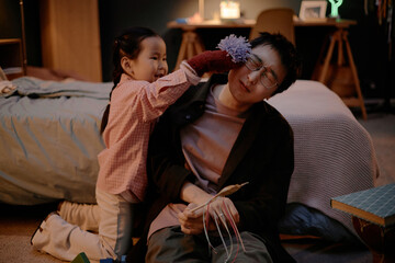 Asian child playfully touching face of Asian young adult man with fuzzy toy while kneeling on floor in bedroom, man holding handmade craft, both engaging in playful family interaction