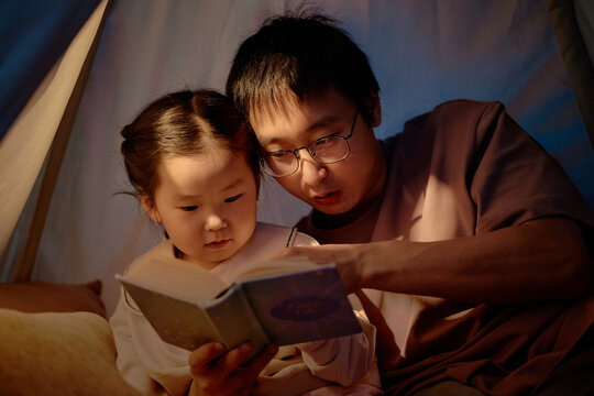 Asian man and child reading book together inside home tent, father guiding daughter through story, both focused on pages, spending evening engaged in family activity