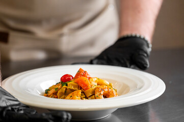 A chef in black gloves presents a white bowl of vibrant, fresh fettuccine gamberi with tomatoes and herbs in a professional kitchen setting, ready to serve