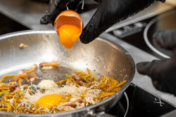 A chef adds a fresh egg yolk to a creamy carbonara pasta dish in a stainless steel pan,...