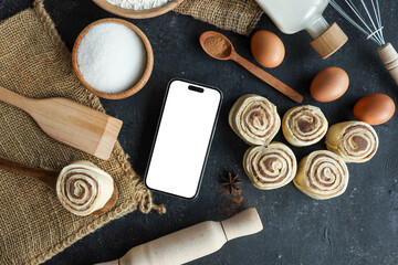 Cinnamon rolls and baking ingredients with a smartphone on a dark background