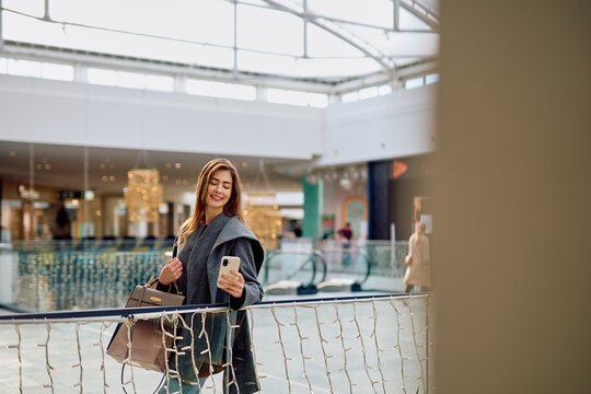 Young woman smiling and holding a smartphone while leaning on a railing in a modern shopping mall, carrying a handbag
