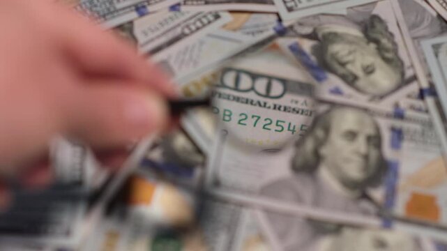 A hand holds a magnifying glass to closely examine the intricate designs of hundred dollar bills placed on a flat surface.