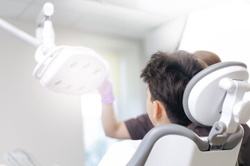 Young woman stomatologist examining teeth of caucasian boy during appointment at dentist office, prepare lighting equipment