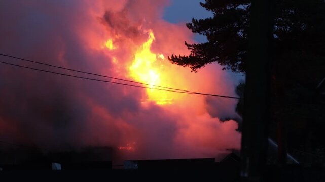 A significant house fire erupts at night, showing bright flames and thick smoke billowing against the dark sky, alarming nearby residents.