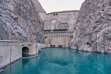 Massive concrete Toktogul HPP dam in a narrow rocky canyon. Turquoise water below, tunnel on the left. High stone walls, clear sky.