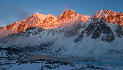 Golden sunrise light illuminating majestic snow-covered mountain peaks over a frozen alpine lake in a serene wilderness