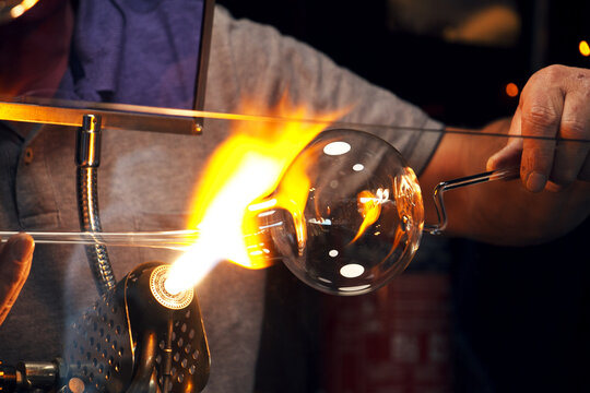 Close-up of a glassblower shaping a clear glass ornament over an open flame. Bright fire and reflections highlight the craftsmanship and traditional handmade production.