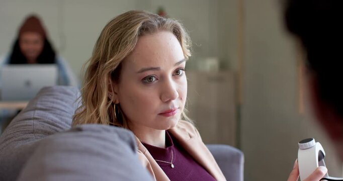 woman in her 20s reacting to coworker talking on sofa holding device, digesting info, smiling