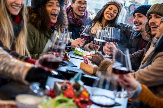 Happy multiracial friends toasting red wine at restaurant terrace - Group of young people wearing winter clothes having fun at outdoors winebar table - Dining life style and friendship concept