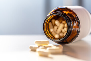 Close view of open amber bottle with blank label and scattered capsules on white surface; selective focus and airy tones support vitamin health messages and minimalist branding.