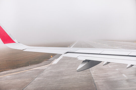 Airplane wing viewed from passenger window as aircraft operates on runway in dense fog. Concrete runway and limited visibility create moody sense of atmosphere during landing or takeoff sequence