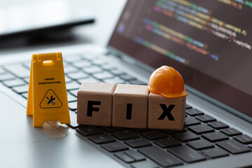 Toy hard hat and "FIX" block on laptop with "Under Construction" sign and code, symbolizing system repair and upgrades