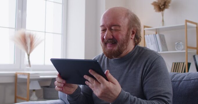 Cheerful bearded elderly man sitting on sofa and holding digital tablet while talking with relatives or friends. Smiling senior male waving hand and enjoying video call or online conversation.
