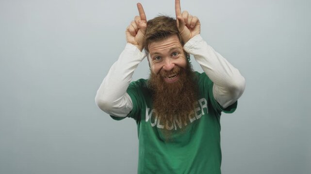 Man with long beard wearing green volunteer shirt makes rock horns gesture on head in studio; fun spirit.