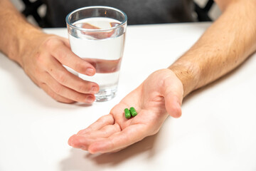 Close-up of male hands holding two green herbal capsules next to a glass of water. Concept of taking daily medication or supplements.