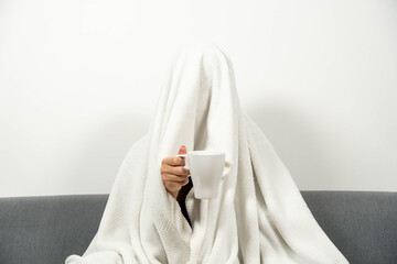 Person completely covered by a white blanket sitting on a sofa holding a hot drink. Concept of flu, winter cold, social anxiety, or Monday mood humor.